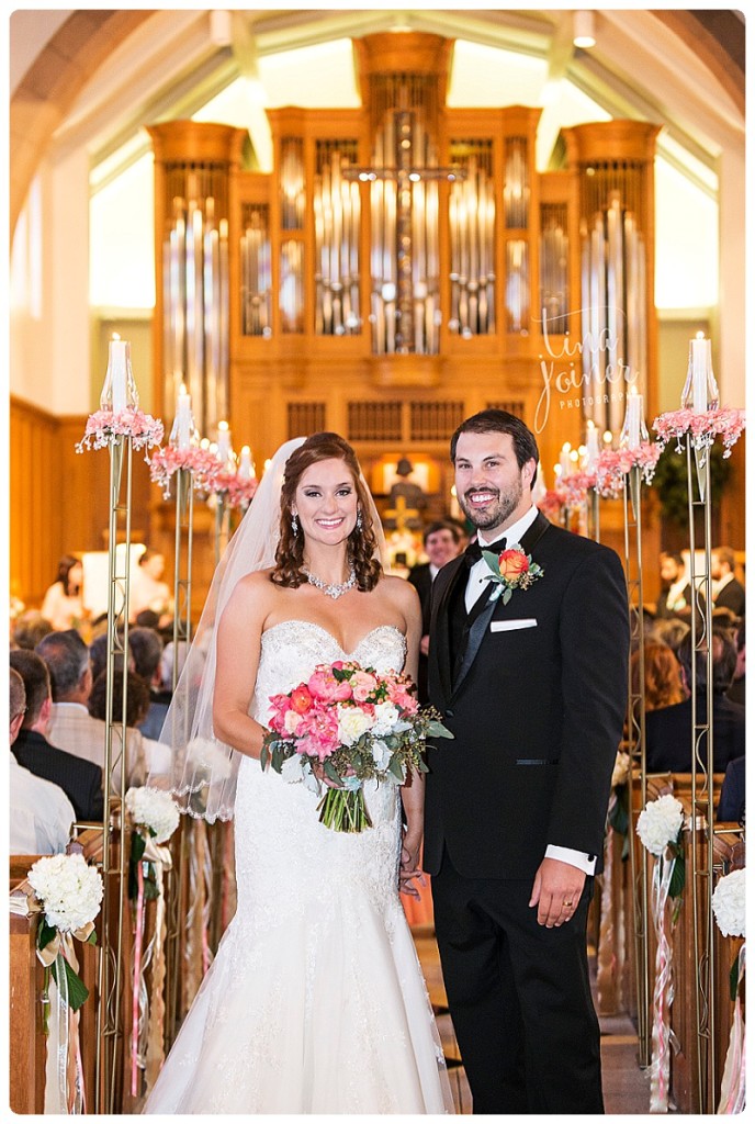 A bride with red hair and a white mermaid wedding dress stands next to her new husband in his black tux, they stand at the end of a church aisle, behind them in the background is a large pipe organ, guests are still sitting in the pews
