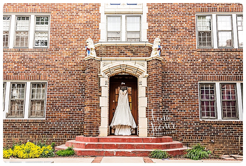 A white, mermaid-style wedding dress hangs on an ornate hanger on the outside of a church door, the building is all brick and there are several windows on the building, there are red steps leading up to the doorway, which has a large white stone frame