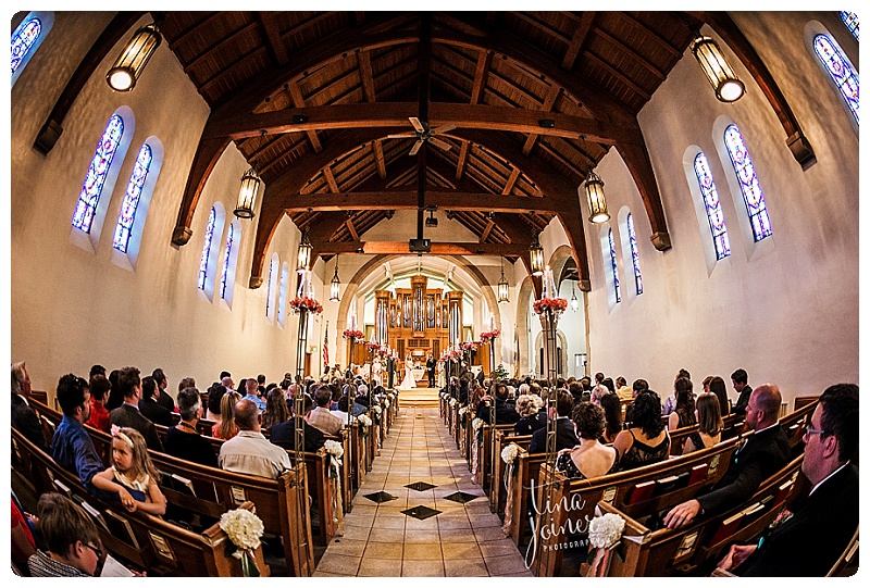 a bride and groom stand near the center of the image at the end of a long aisle during their wedding ceremony, the image was taken with a fisheye lens, the ceiling of the church sanctuary his wood and has arches, the pews are also wood and line the walls, which have pairs of stain glass windows along them, there is a large pipe organ on the altar stage