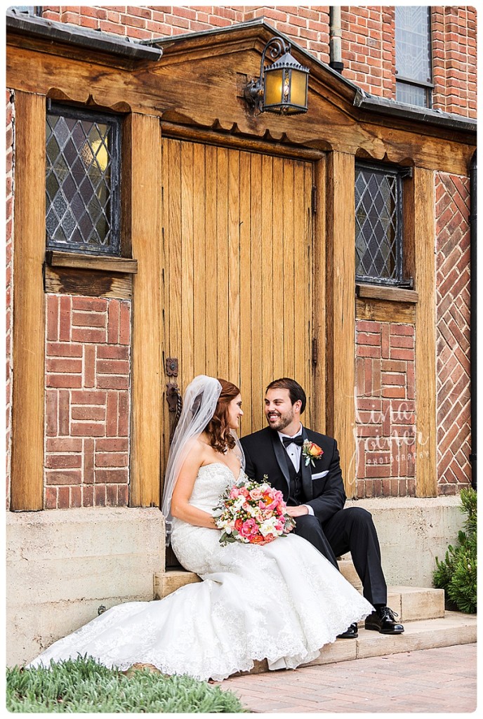 A bride and groom sit together on steps in front of a brick building with a wood door, she holds her pink bouquet in her hands, they look at each other and smile, the groom wears a black bow tie with his tux