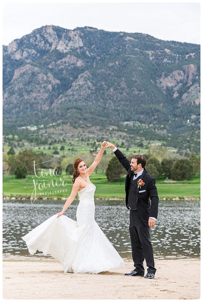 A groom in a black tux and black bow tie twirls his bride in a white, strapless, mermaid wedding dress, they stand on sand with a body of water, golf course, and mountain behind them, the groom looks at the bride and the bride looks at the camera as she spins