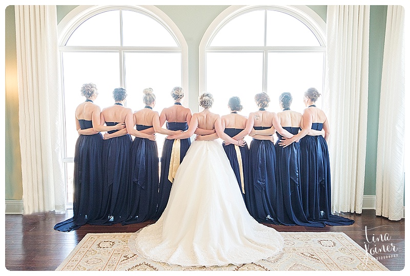 The backs of a bride and 8 bridesmaids standing with their arms around each other at The Pinery at the Hill in the bridal room with two huge windows