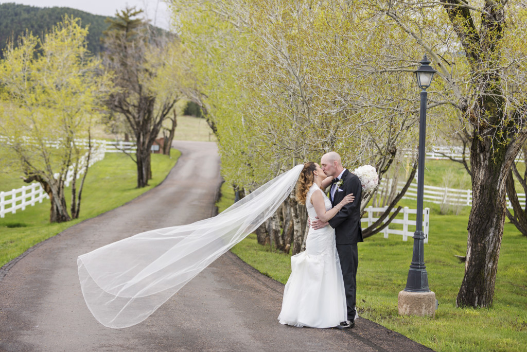 Bride and groom stand and embrace while kissing, brides veil flies behind her