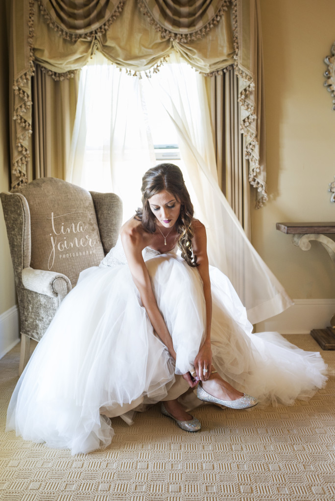 Bride sits in a chair by a window and straps her shoe on
