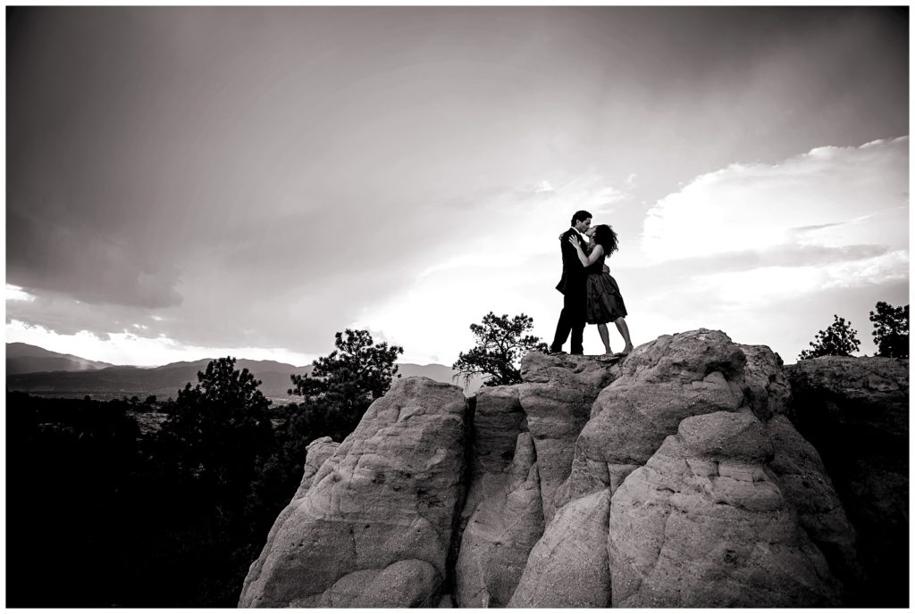 Samuel and Pam share a kiss on large rocks at Palmer Park in black and white, behind them are stunning mountain views and a clear sky