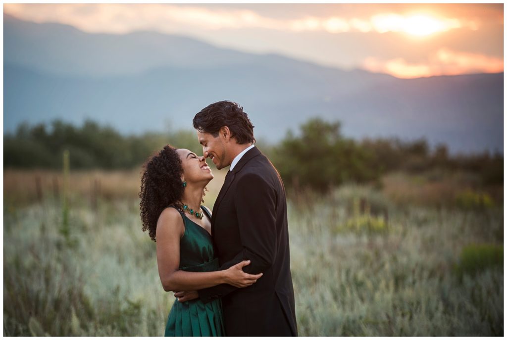 Samuel and Pam are laughing and giving each other an eskimo kiss, they are embracing, they are standing in a field with mountains and sunset behind them