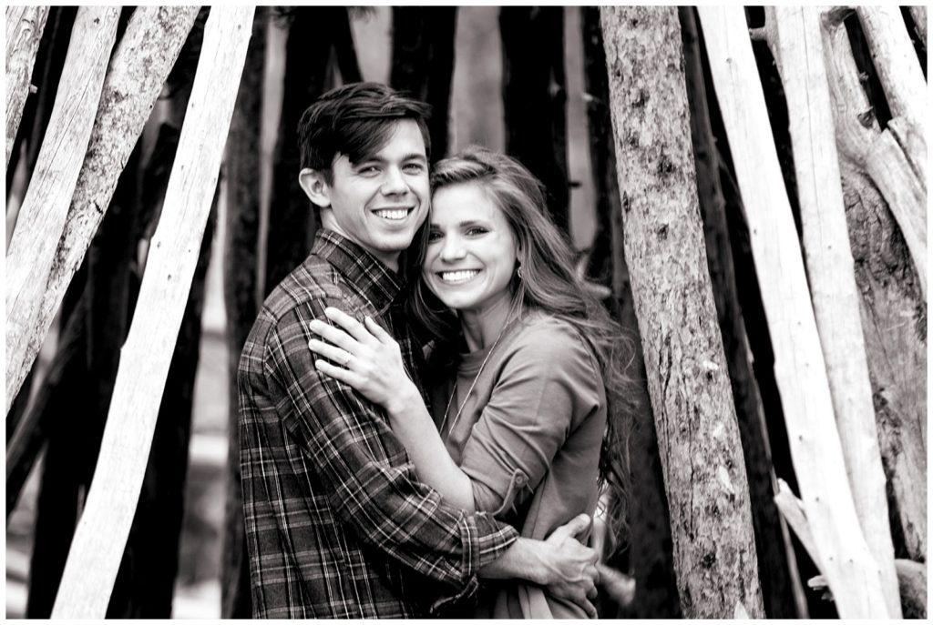 Stephanie and Tate embrace under an old fort built of logs in black and white, they are looking at the camera and smiling