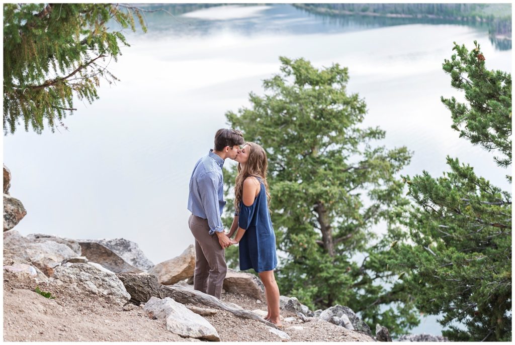 Stephanie and Tate are kissing each other while holding hands standing at the top of Sapphire Point, behind them is a lake