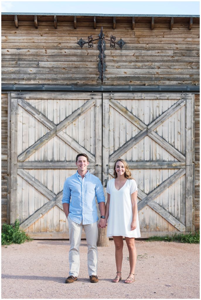 Conner and Megan are holding hands and standing in front of the doors of an old brown barn, this is a front perspective and the couple is smiling and facing forward