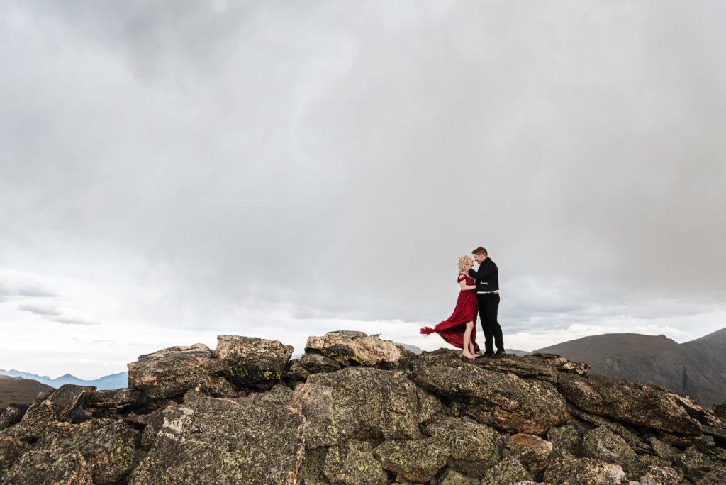 Kurtis and Mollie stand on a rocky mountain top at Rocky Mountain National Park, the wind is moving her long red dress, they are embracing and looking at each other smiling