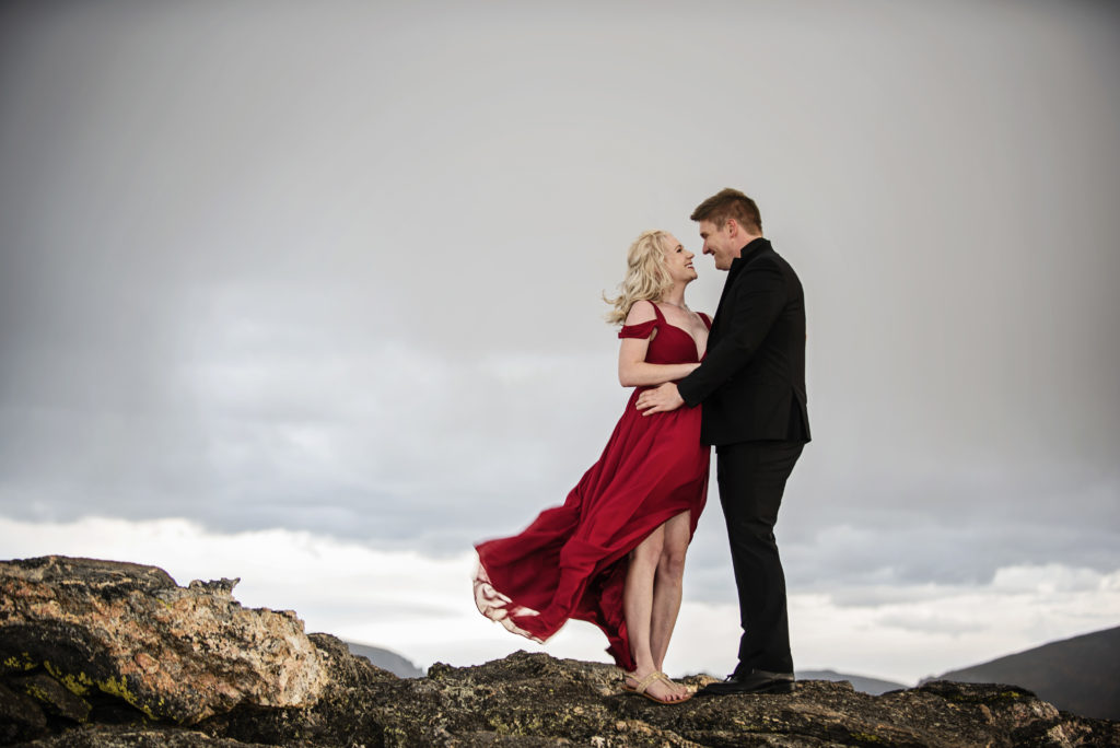 Kurtis and Mollie stand on a windy mountain top looking at each other and smiling, she is wearing a long red dress that is being whipped by the wind