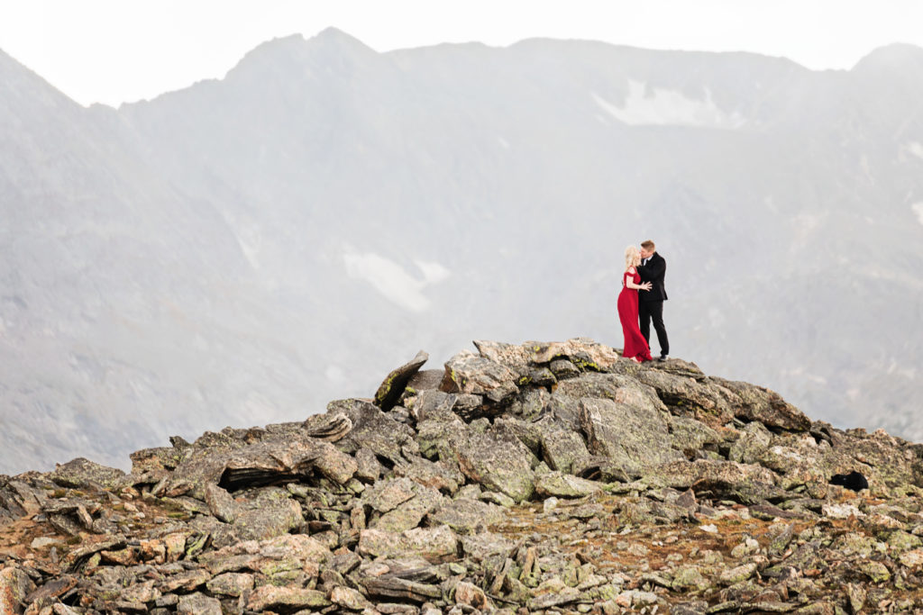 Couple wearing formal outfits stands on a mountain top with grand mountain views embracing and kissing, she is in a long red dress, and he is wearing a black suit