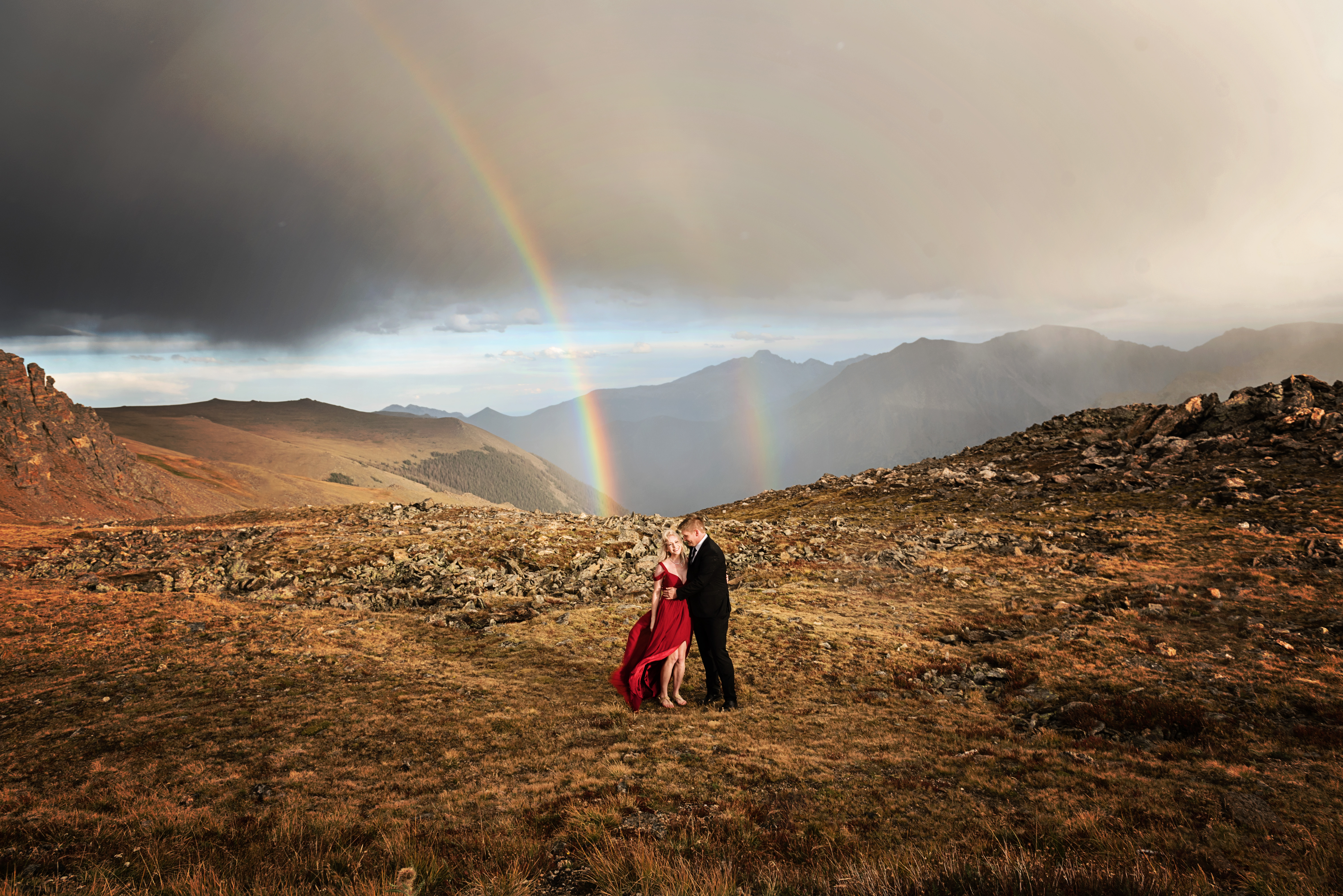 Couple stand on rocky mountain top at Rocky Mountain National Park looking at each other with double rainbow and grand mountain views behind them.