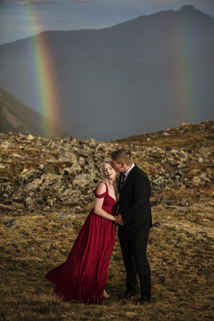 Kurtis and Mollie are holding hands and cuddling on a mountain top, he is kissing her temple and she has her eyes closed as wind sweeps through her hair and dress, behind them is a rainbow