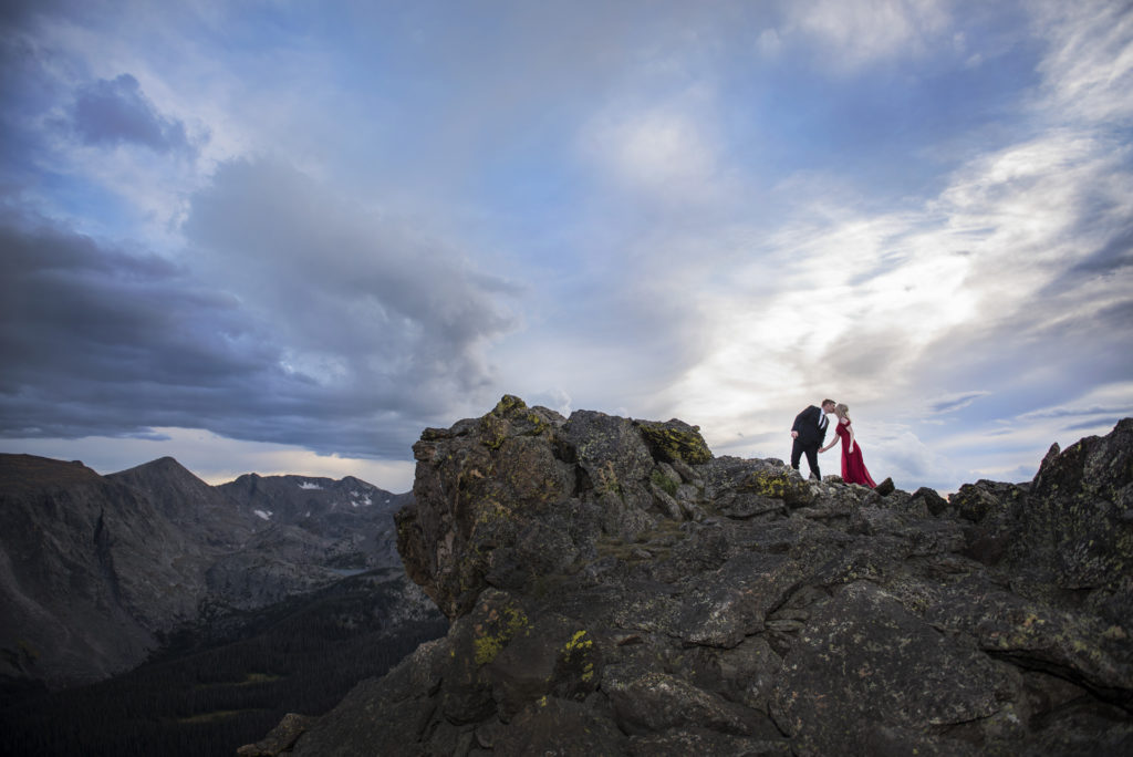 Curtis and Mollie are kissing on a large rock with a wide open mountain view and amazing blue sky with thick white clouds, she is in a red dress and he is wearing a black suit