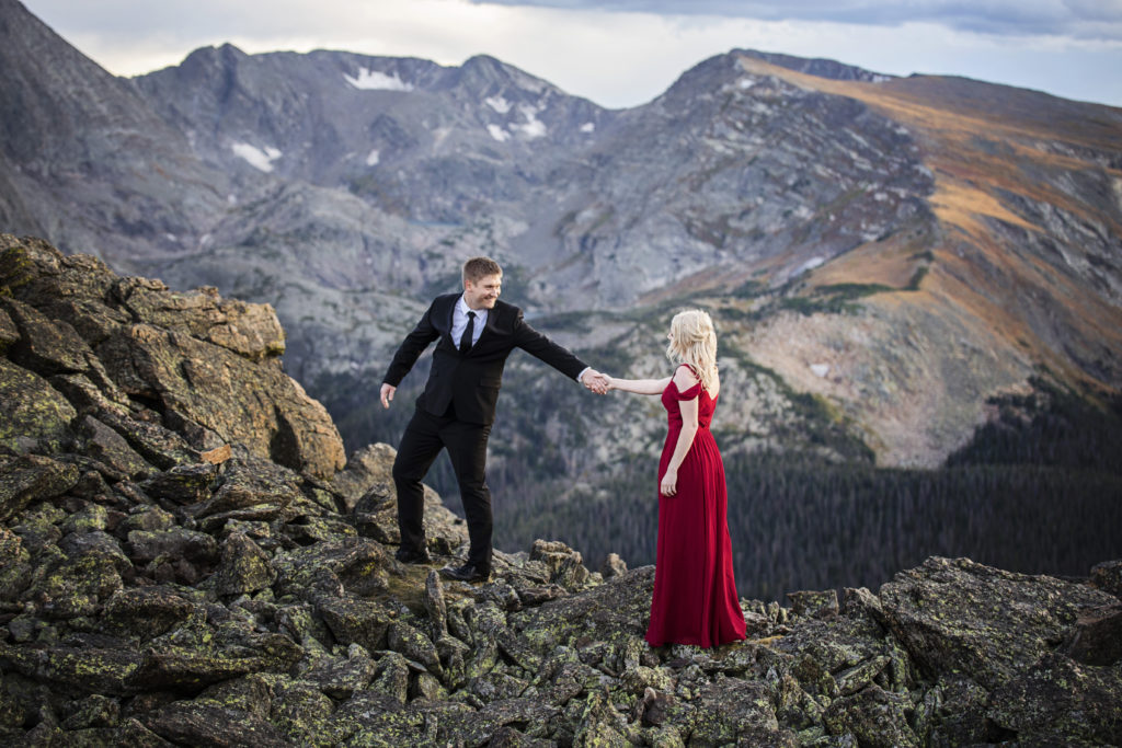 Kurtis is leading Mollie by the hand on a mountain top at Rocky Mountain National Park, behind them is an amazing view of the mountains, they are in formal wear