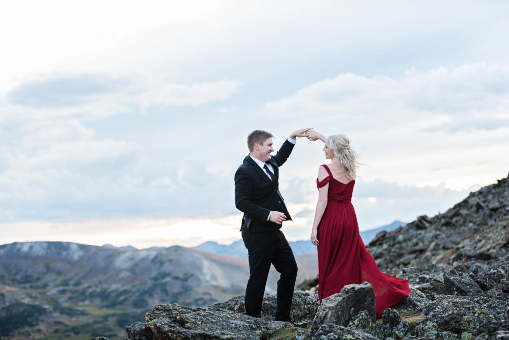 Kurtis and Mollie dance on a mountain top, she is wearing a long read gown and he is wearing a black suit