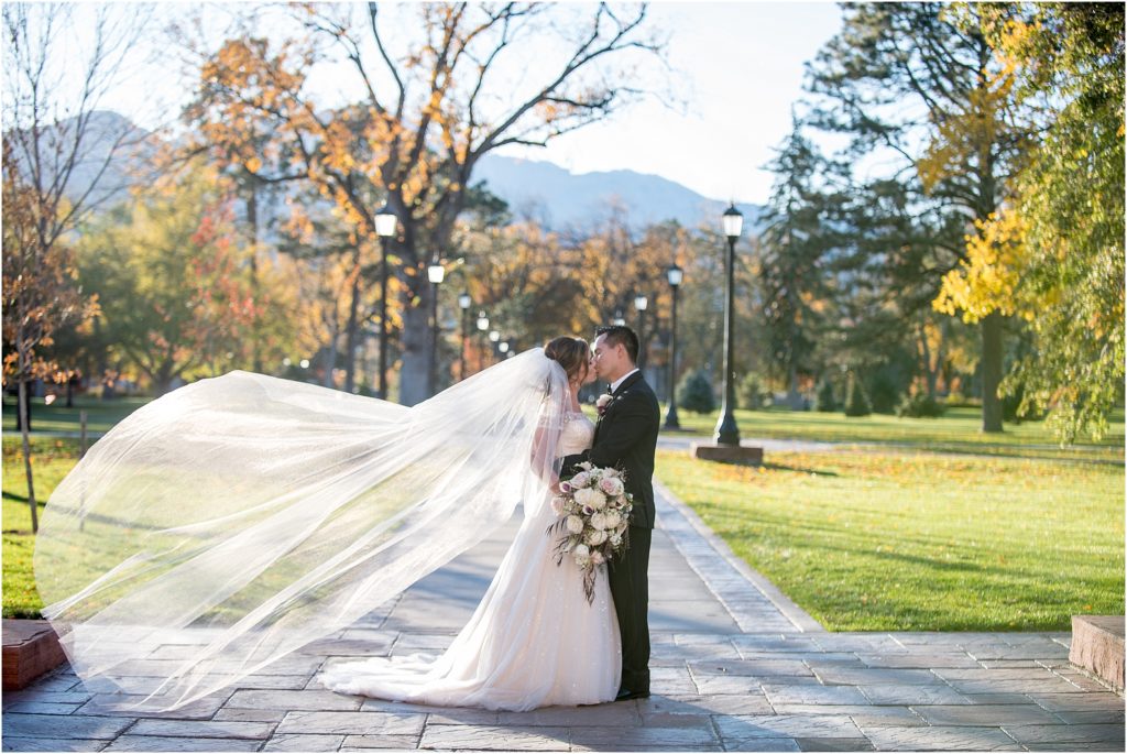 Bride and groom stand on manicured lawn embracing and kissing as her veil flies behind her in the fall