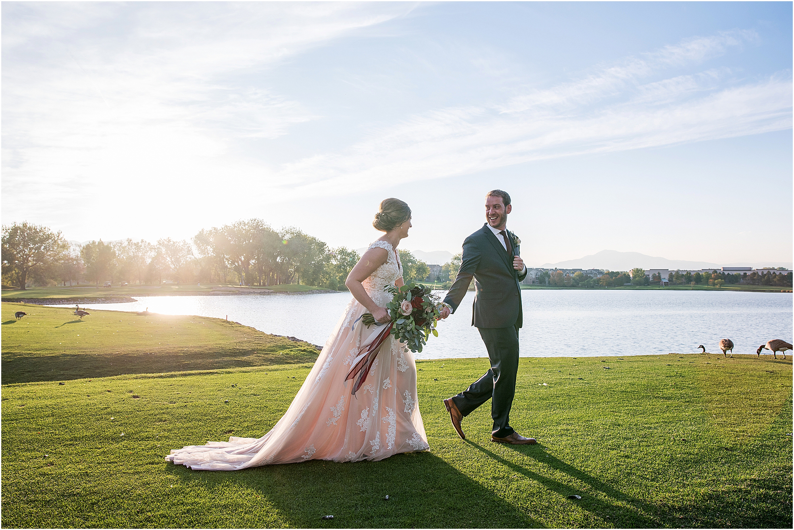 taken at sunset, bride and groom walk holding hands, groom leads bride, bride is holding bouquet and smiling at the groom, there are geese and a pond