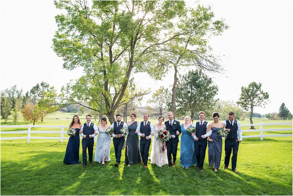 Bride and groom walk with their wedding party in a field with a white fence behind them