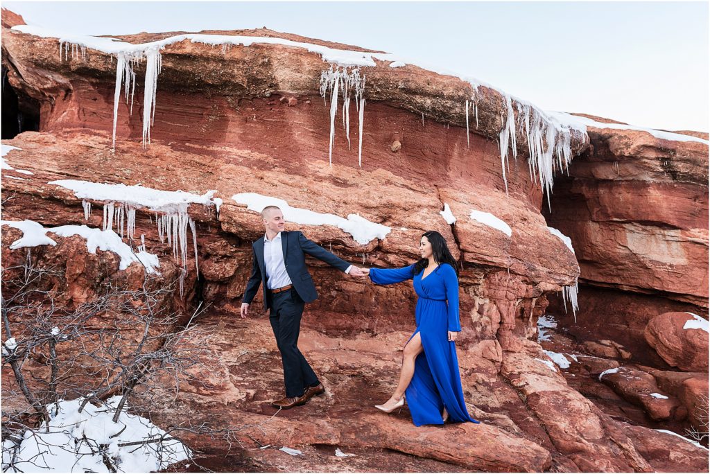 Tom leads Celene on a rock at Garden of the Gods in winter, they are holding hands and smiling wearing formal clothes