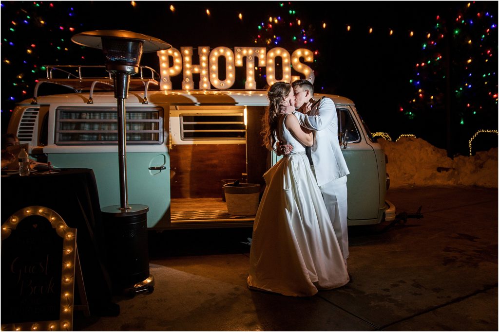 Bride and groom stand embracing and kissing in front of the Shutterbus photo booth at night at Flying Horse Ranch