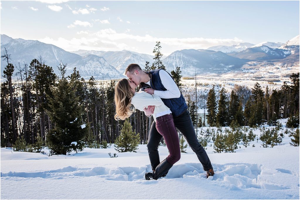 Man dips woman on top of a mountain in winter, they are kissing, mountain views and forested trees behind them