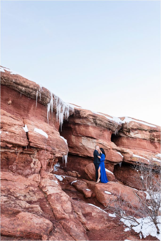 A romantic couple is embracing at Garden of the Gods in winter, she is wearing a blue gown and he is wearing a suit