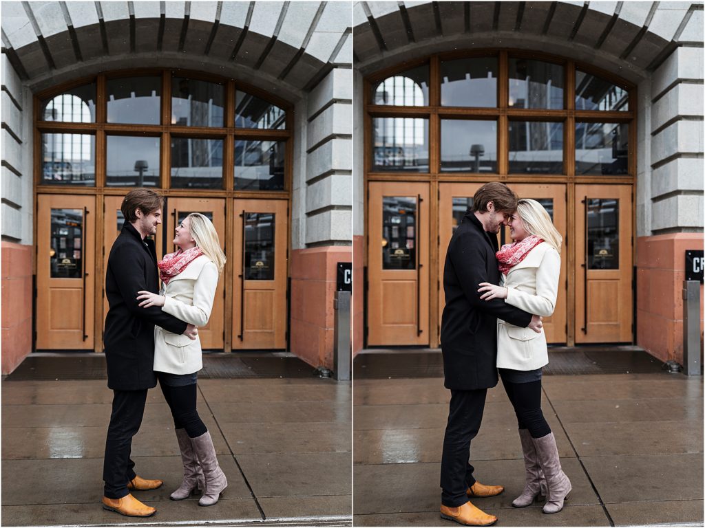 Engaged couple stand and embrace and look at each other with smiles in front of beautiful wooden doors with glass windows