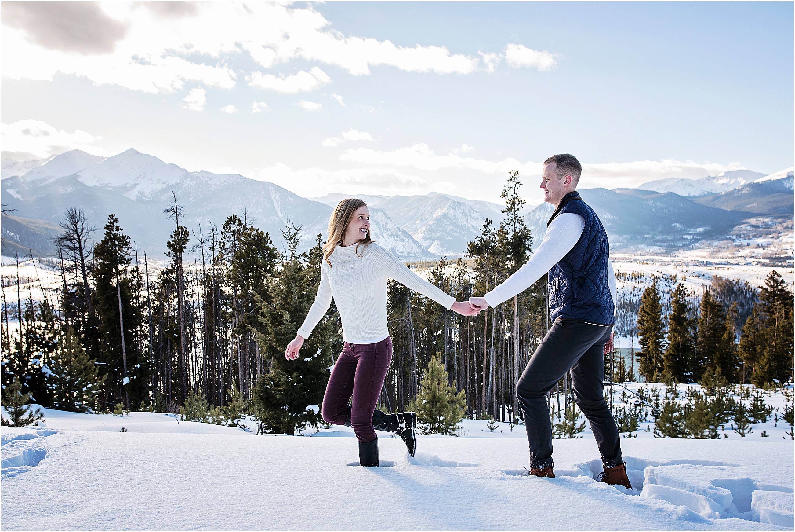 Young couple walk in the snow with the woman in front looking back at her fiancé, they are holding hands and smiling, stunning mountain views behind them, near Breckenridge Colorado