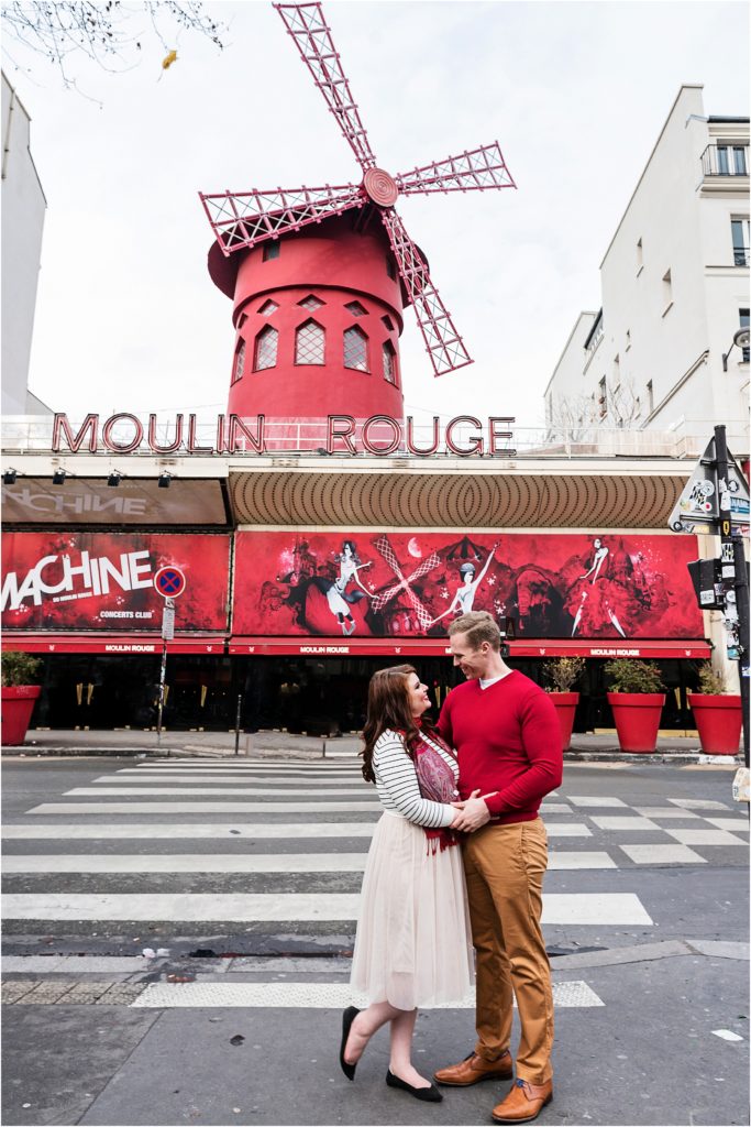 Couple standing and embracing smiling at each other with the Moulin Rouge behind them in Paris