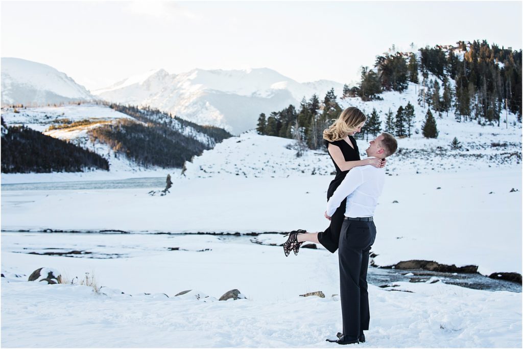Man lifts woman and they look at each other with snow all around and mountain views and a frozen pond behind them