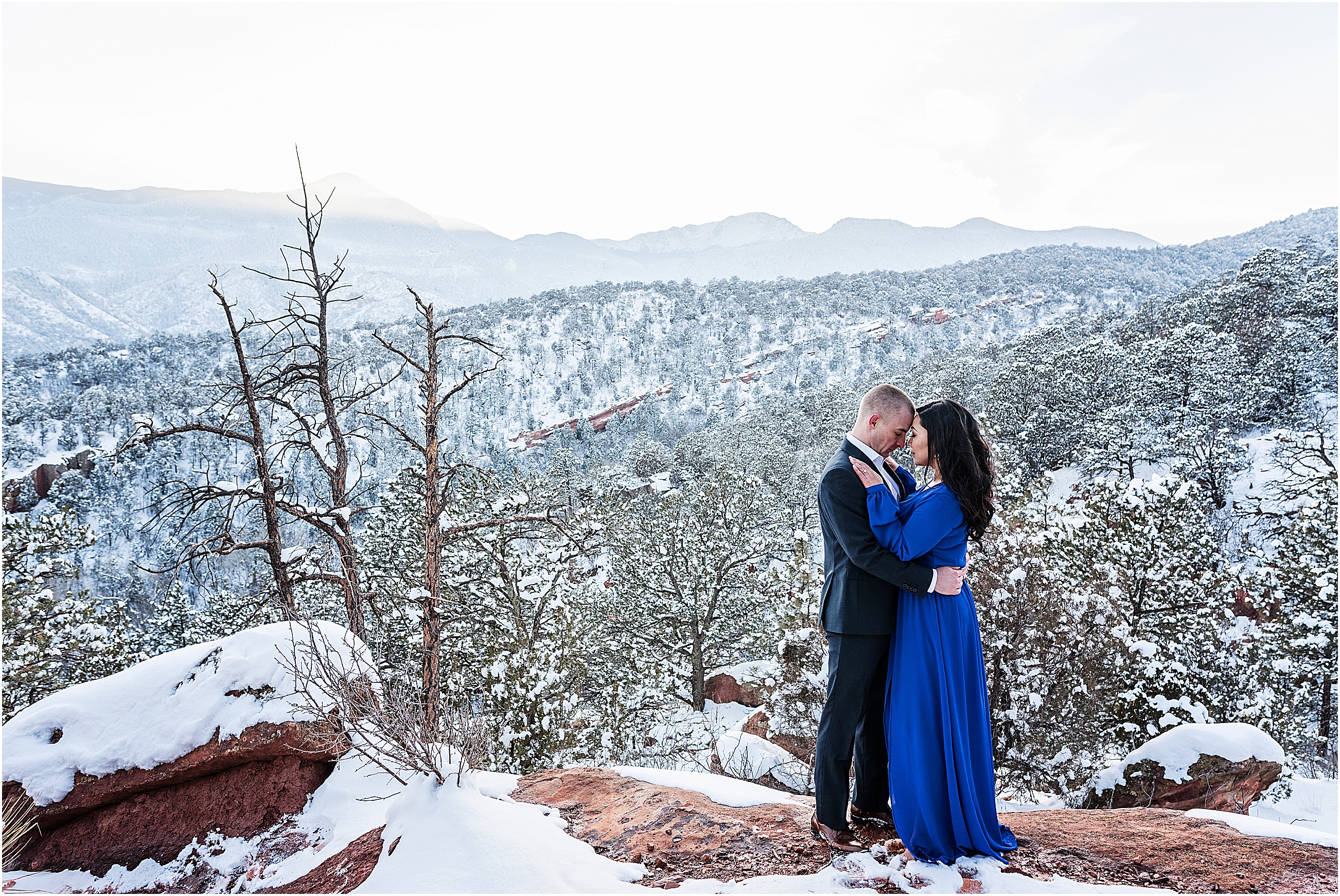 A couple stands holding each other wearing formal clothes overlooking a winter wonderland with snow and ice all round and mountain views behind