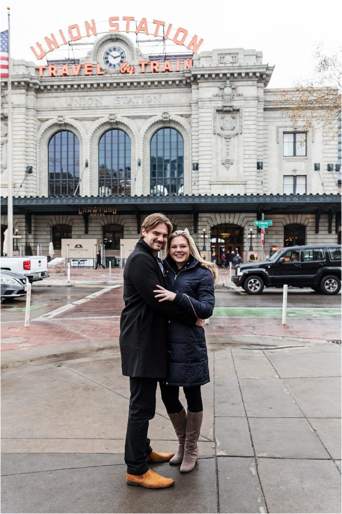Engaged couple are standing and embracing and smiling while union station is in the background and snow falls around them