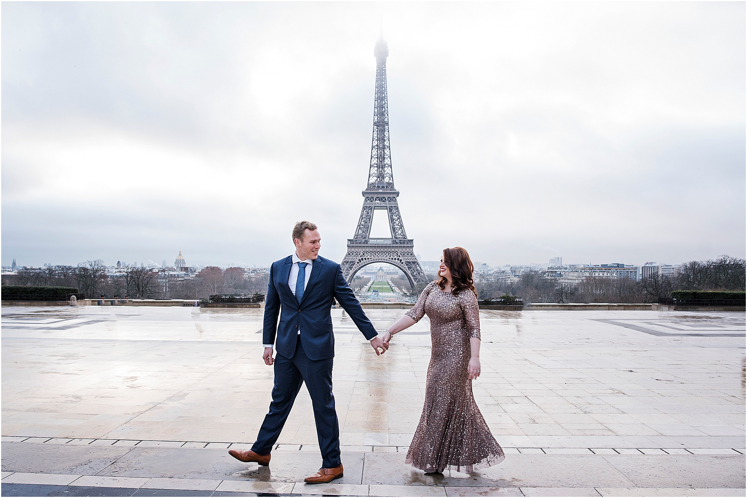 Engaged couple walk holding hands looking at each other and smiling with the Eiffel Tower behind them in Paris
