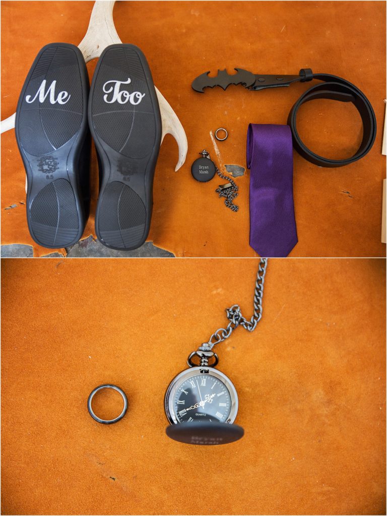 Lay flat of grooms details that include an engraved pocket watch, his wedding band, dark purple tie, batman belt, and his shoes that have "me too" on the bottom of them
