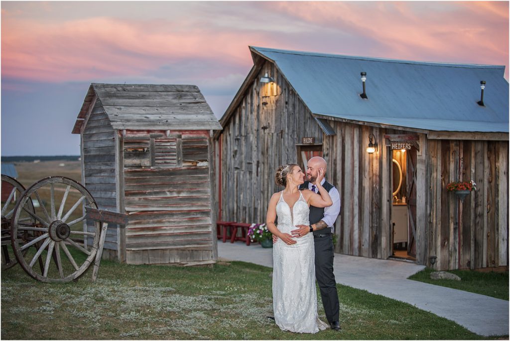 Bryan embraces Lori from behind and she reaches to touch his cheek, they stand in front of the bridal cottage at Younger Ranch in Colorado at sunset, the sky is pink and blue and purple