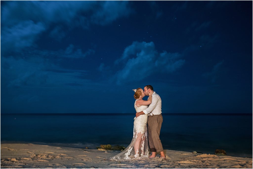 bride and groom embrace and kiss by the ocean at night, the stars can be seen between the clouds