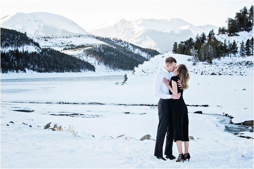Couple stands in formal clothes, embracing and kissing with snowy mountain views and a frozen pond behind them