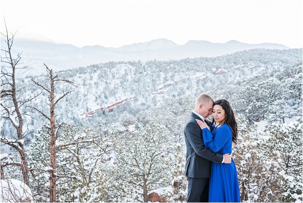 Tom and Celene hold each other, Tom has his forehead on her temple and both have their eyes closed, they are wearing formal clothes at Garden of the Gods in winter, snow is all around them