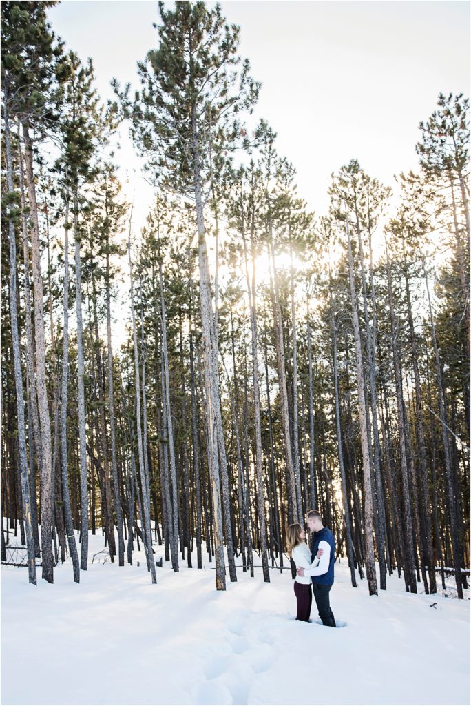 Young couple stand in snow to their knees, they look at each other while they embrace in a forest where the trees stretch high into the sky