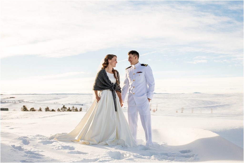 bride and groom hold hands and look at each other and smile while standing in fresh snow in a field at their winter wedding