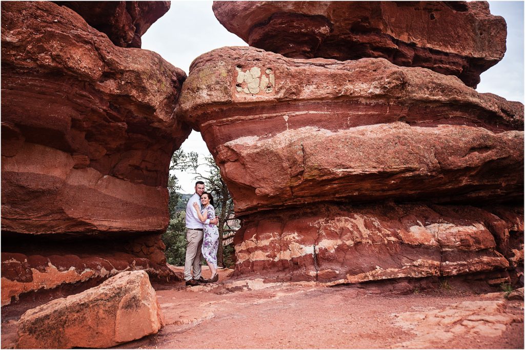 Engaged man and woman stand embracing between two very large red rocks at Garden of the Gods in Colorado Springs