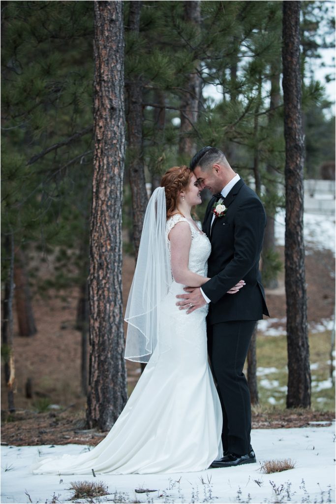 Bride and groom stand in snow in a forest with their foreheads together after their wedding ceremony and smile