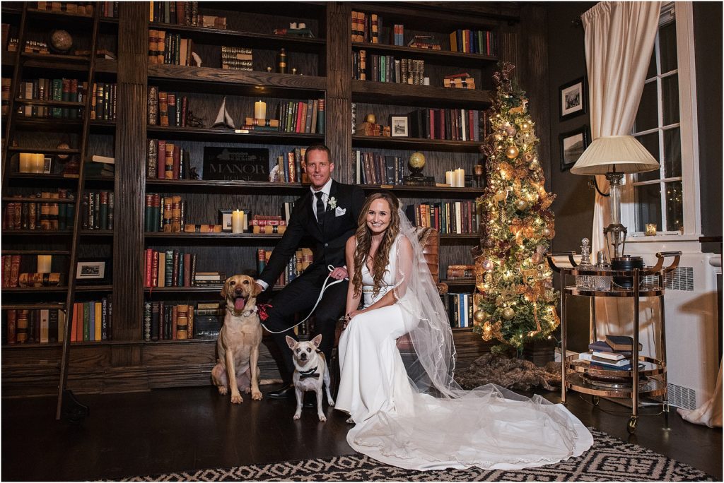 Bride and groom are sitting with their two dogs in the library at their Holiday wedding at The Manor House