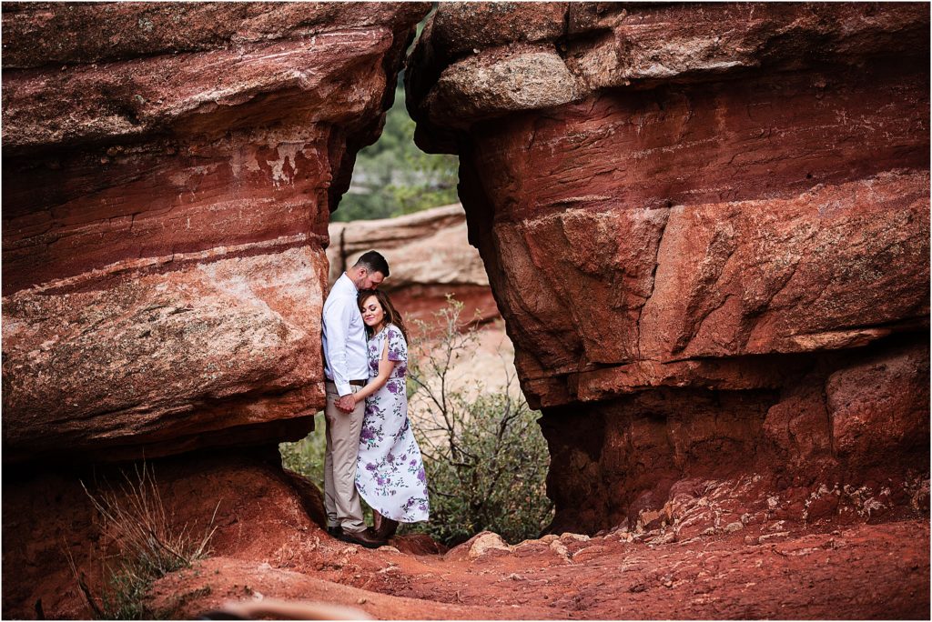 Man romantically kissing woman on the head as they embrace between large red boulders at Garden of the Gods
