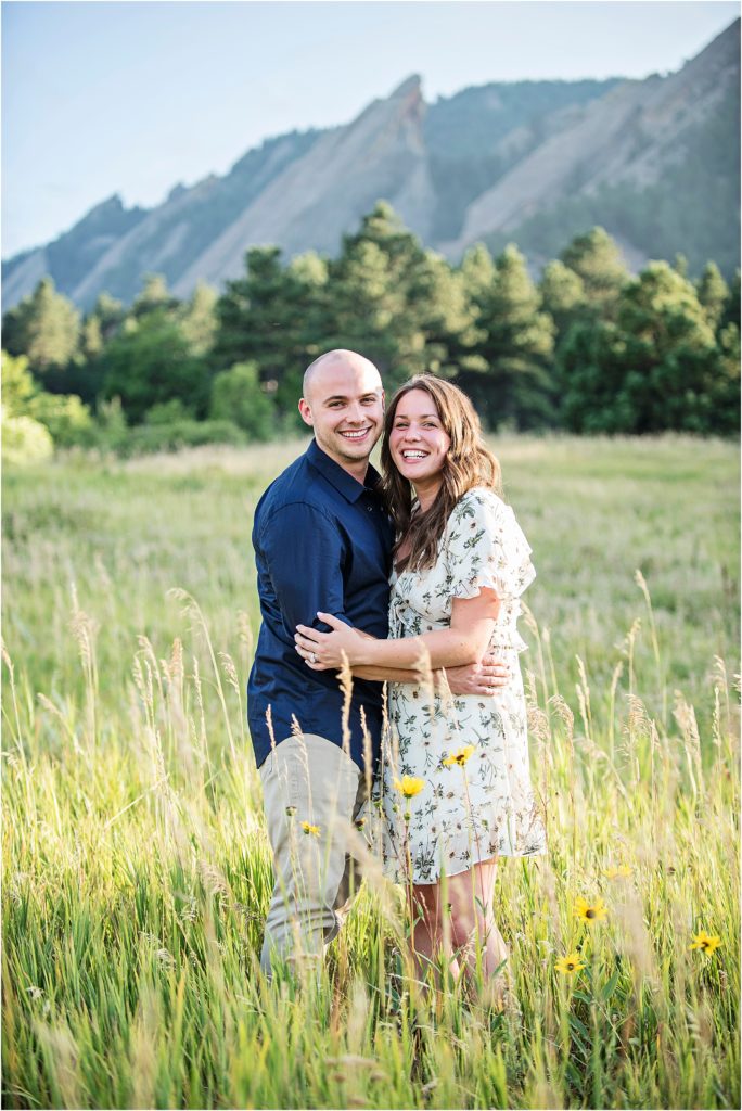Engaged couple stand and embrace and smile with Chautauqua Flatirons behind them in summer