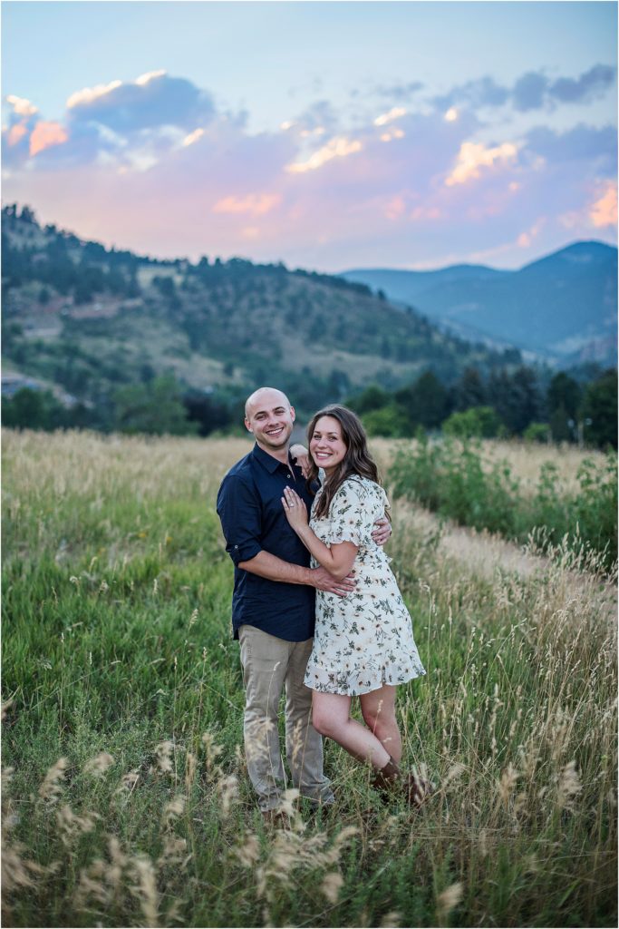 Engaged couple stand in meadow at sunset with mountains and a blue, pink, and purple sky behind them.