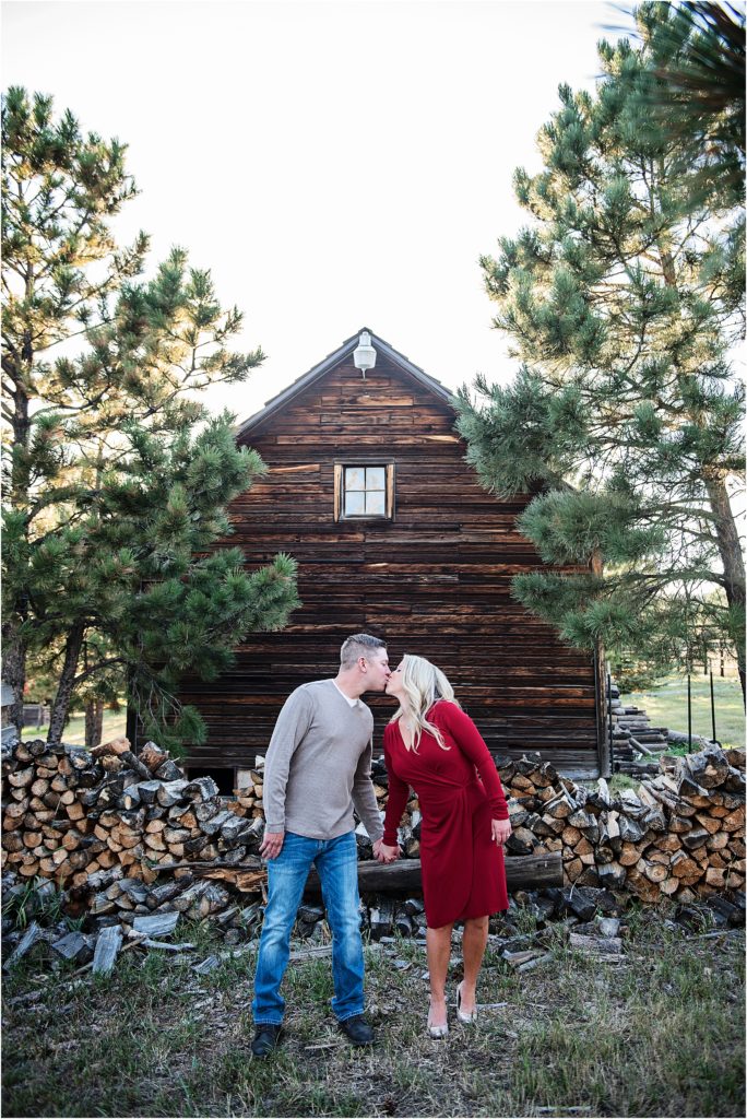 Brian and Jordan share a kiss in front of a wood pile and a rustic cabin at Spruce Mountain Ranch in Colorado