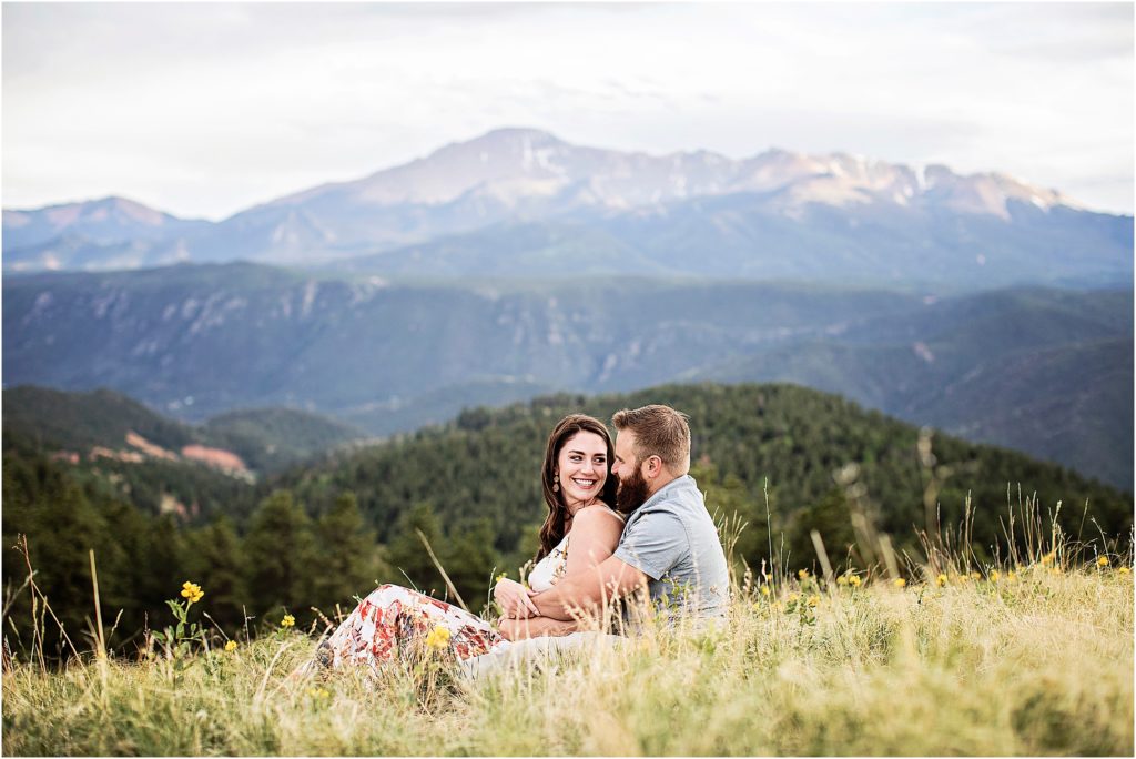 Couple sits and embraces on a hilltop with stunning views of Pikes Peak behind them